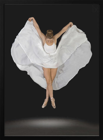 A high-angle studio photograph of a graceful ballet dancer suspended mid-air against a solid black background. She wears a white leotard and pointe shoes, with her legs crossed. Her arms are outstretched, holding a large, flowing white fabric that creates a shape like wings around her. Decor