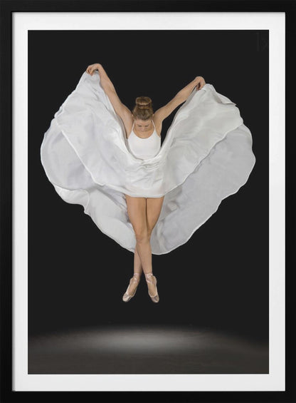 A high-angle studio photograph of a graceful ballet dancer suspended mid-air against a solid black background. She wears a white leotard and pointe shoes, with her legs crossed. Her arms are outstretched, holding a large, flowing white fabric that creates a shape like wings around her. Decor