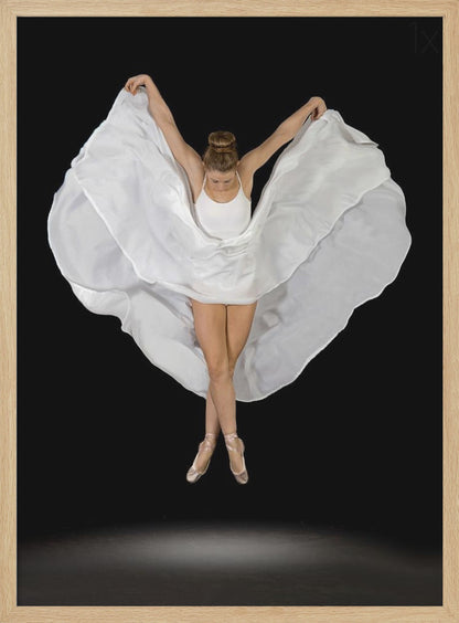 A high-angle studio photograph of a graceful ballet dancer suspended mid-air against a solid black background. She wears a white leotard and pointe shoes, with her legs crossed. Her arms are outstretched, holding a large, flowing white fabric that creates a shape like wings around her. Decor