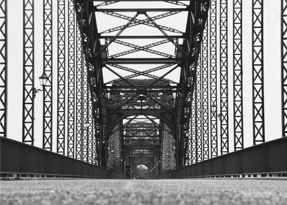 A framed, black and white, low-angle photograph capturing the symmetrical and intricate steel truss structure of a long bridge, creating a powerful vanishing point perspective down the center of the road. Poster