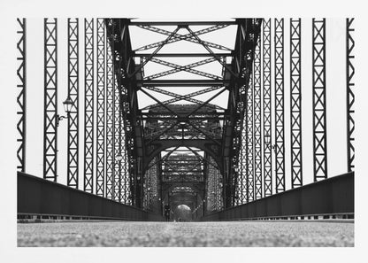 A framed, black and white, low-angle photograph capturing the symmetrical and intricate steel truss structure of a long bridge, creating a powerful vanishing point perspective down the center of the road. Poster