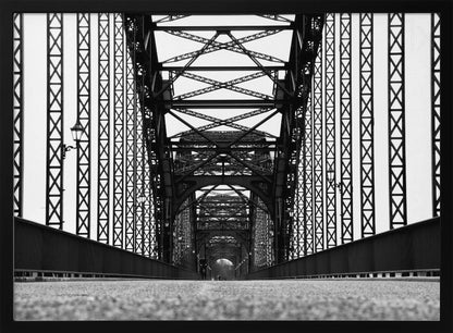 A framed, black and white, low-angle photograph capturing the symmetrical and intricate steel truss structure of a long bridge, creating a powerful vanishing point perspective down the center of the road. Poster