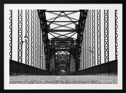 A framed, black and white, low-angle photograph capturing the symmetrical and intricate steel truss structure of a long bridge, creating a powerful vanishing point perspective down the center of the road. Poster