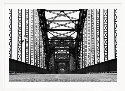A framed, black and white, low-angle photograph capturing the symmetrical and intricate steel truss structure of a long bridge, creating a powerful vanishing point perspective down the center of the road. Poster