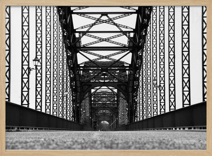 A framed, black and white, low-angle photograph capturing the symmetrical and intricate steel truss structure of a long bridge, creating a powerful vanishing point perspective down the center of the road. Poster