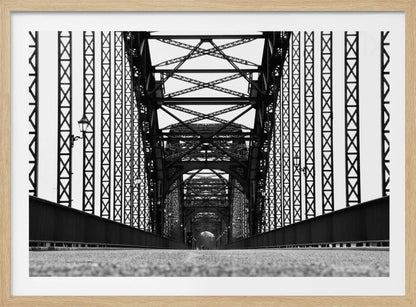 A framed, black and white, low-angle photograph capturing the symmetrical and intricate steel truss structure of a long bridge, creating a powerful vanishing point perspective down the center of the road. Poster