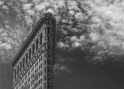 A dramatic low-angle black and white photograph of the Flatiron Building in New York City, with its sharp, triangular corner pointing towards a sky filled with fluffy, white clouds. The image is framed with a brushed silver border. Print
