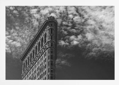 A dramatic low-angle black and white photograph of the Flatiron Building in New York City, with its sharp, triangular corner pointing towards a sky filled with fluffy, white clouds. The image is framed with a brushed silver border. Print