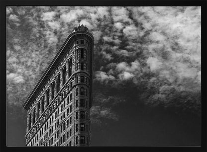 A dramatic low-angle black and white photograph of the Flatiron Building in New York City, with its sharp, triangular corner pointing towards a sky filled with fluffy, white clouds. The image is framed with a brushed silver border. Print