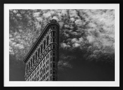 A dramatic low-angle black and white photograph of the Flatiron Building in New York City, with its sharp, triangular corner pointing towards a sky filled with fluffy, white clouds. The image is framed with a brushed silver border. Print