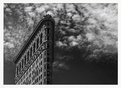 A dramatic low-angle black and white photograph of the Flatiron Building in New York City, with its sharp, triangular corner pointing towards a sky filled with fluffy, white clouds. The image is framed with a brushed silver border. Print