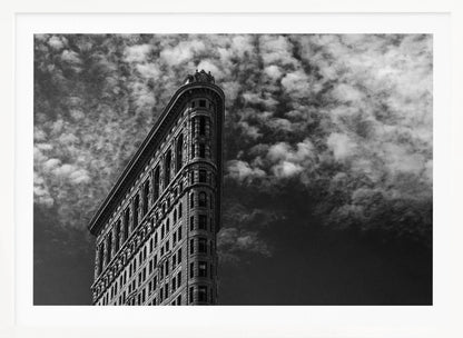 A dramatic low-angle black and white photograph of the Flatiron Building in New York City, with its sharp, triangular corner pointing towards a sky filled with fluffy, white clouds. The image is framed with a brushed silver border. Print