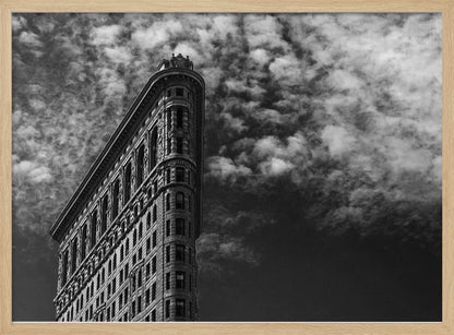 A dramatic low-angle black and white photograph of the Flatiron Building in New York City, with its sharp, triangular corner pointing towards a sky filled with fluffy, white clouds. The image is framed with a brushed silver border. Print