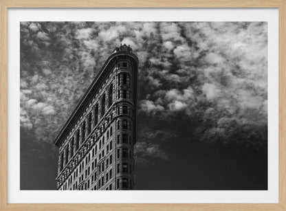A dramatic low-angle black and white photograph of the Flatiron Building in New York City, with its sharp, triangular corner pointing towards a sky filled with fluffy, white clouds. The image is framed with a brushed silver border. Print