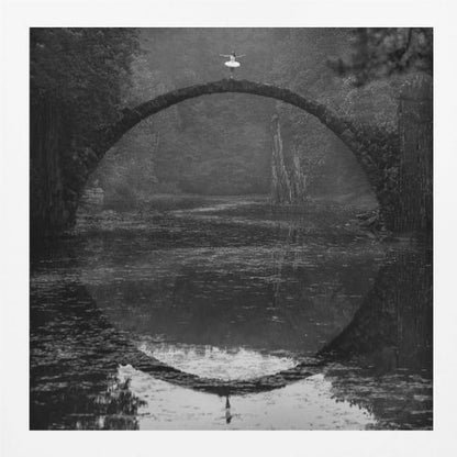 A framed black and white photograph of a ballerina in a white tutu standing at the peak of a semi-circular stone bridge. The bridge and the ballerina are perfectly reflected in the still water below, creating a full circle. The scene is set in a dark, misty forest, giving it a surreal and moody atmosphere. Artwork
