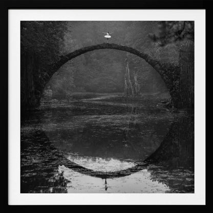 A framed black and white photograph of a ballerina in a white tutu standing at the peak of a semi-circular stone bridge. The bridge and the ballerina are perfectly reflected in the still water below, creating a full circle. The scene is set in a dark, misty forest, giving it a surreal and moody atmosphere. Artwork