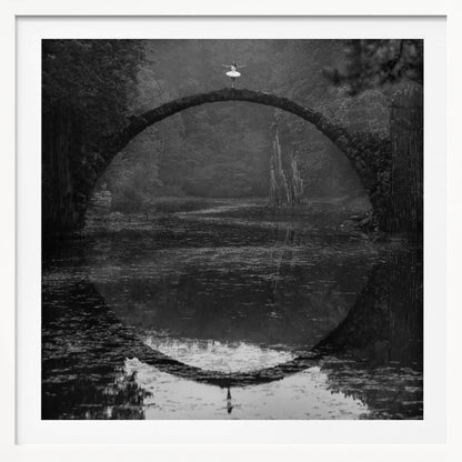 A framed black and white photograph of a ballerina in a white tutu standing at the peak of a semi-circular stone bridge. The bridge and the ballerina are perfectly reflected in the still water below, creating a full circle. The scene is set in a dark, misty forest, giving it a surreal and moody atmosphere. Artwork