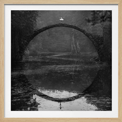 A framed black and white photograph of a ballerina in a white tutu standing at the peak of a semi-circular stone bridge. The bridge and the ballerina are perfectly reflected in the still water below, creating a full circle. The scene is set in a dark, misty forest, giving it a surreal and moody atmosphere. Artwork