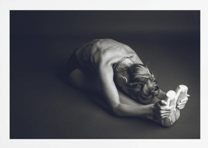 A black and white photograph of a ballerina stretching. She is seated on the floor in a deep forward fold, her torso bent over her legs as she holds onto her pointe shoes. The lighting accentuates the musculature of her back and shoulders, and her hair is styled in a braided bun. Decor