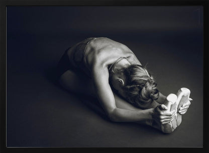 A black and white photograph of a ballerina stretching. She is seated on the floor in a deep forward fold, her torso bent over her legs as she holds onto her pointe shoes. The lighting accentuates the musculature of her back and shoulders, and her hair is styled in a braided bun. Decor
