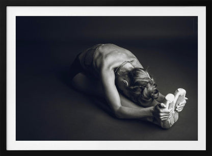 A black and white photograph of a ballerina stretching. She is seated on the floor in a deep forward fold, her torso bent over her legs as she holds onto her pointe shoes. The lighting accentuates the musculature of her back and shoulders, and her hair is styled in a braided bun. Decor
