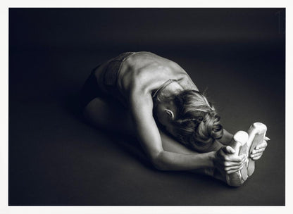 A black and white photograph of a ballerina stretching. She is seated on the floor in a deep forward fold, her torso bent over her legs as she holds onto her pointe shoes. The lighting accentuates the musculature of her back and shoulders, and her hair is styled in a braided bun. Decor