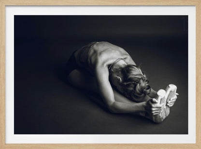 A black and white photograph of a ballerina stretching. She is seated on the floor in a deep forward fold, her torso bent over her legs as she holds onto her pointe shoes. The lighting accentuates the musculature of her back and shoulders, and her hair is styled in a braided bun. Decor
