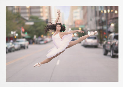 A ballerina in a white tutu and pointe shoes leaps into a split mid-air in the middle of a blurred city street, her arms extended gracefully. The image is set within a silver frame. Print