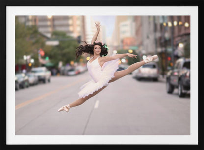 A ballerina in a white tutu and pointe shoes leaps into a split mid-air in the middle of a blurred city street, her arms extended gracefully. The image is set within a silver frame. Print