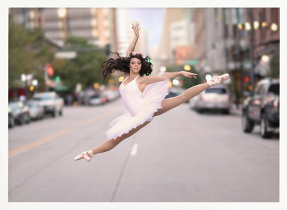 A ballerina in a white tutu and pointe shoes leaps into a split mid-air in the middle of a blurred city street, her arms extended gracefully. The image is set within a silver frame. Print