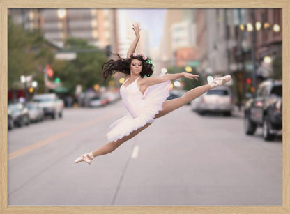A ballerina in a white tutu and pointe shoes leaps into a split mid-air in the middle of a blurred city street, her arms extended gracefully. The image is set within a silver frame. Print