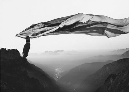 A dramatic black and white photograph of a woman standing on the edge of a mountain peak. She holds up a massive piece of dark, sheer fabric that billows in the wind, stretching across the sky. Below her, a vast, misty mountain range and a deep valley with city lights are visible. Wall Art