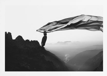 A dramatic black and white photograph of a woman standing on the edge of a mountain peak. She holds up a massive piece of dark, sheer fabric that billows in the wind, stretching across the sky. Below her, a vast, misty mountain range and a deep valley with city lights are visible. Wall Art