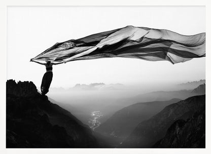 A dramatic black and white photograph of a woman standing on the edge of a mountain peak. She holds up a massive piece of dark, sheer fabric that billows in the wind, stretching across the sky. Below her, a vast, misty mountain range and a deep valley with city lights are visible. Wall Art