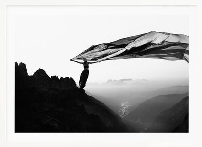 A dramatic black and white photograph of a woman standing on the edge of a mountain peak. She holds up a massive piece of dark, sheer fabric that billows in the wind, stretching across the sky. Below her, a vast, misty mountain range and a deep valley with city lights are visible. Wall Art