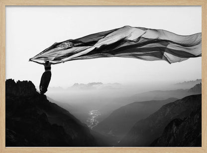 A dramatic black and white photograph of a woman standing on the edge of a mountain peak. She holds up a massive piece of dark, sheer fabric that billows in the wind, stretching across the sky. Below her, a vast, misty mountain range and a deep valley with city lights are visible. Wall Art