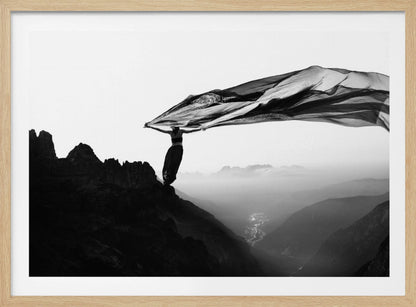 A dramatic black and white photograph of a woman standing on the edge of a mountain peak. She holds up a massive piece of dark, sheer fabric that billows in the wind, stretching across the sky. Below her, a vast, misty mountain range and a deep valley with city lights are visible. Wall Art