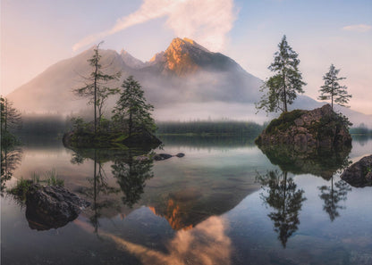 A serene mountain lake at sunrise or sunset, with a prominent peak illuminated by golden light. Small, rocky islands with pine trees dot the calm, clear water, which perfectly reflects the sky, trees, and mountains. A light mist hangs over the water and around the base of the mountains in the background. Decor