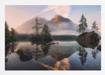 A serene mountain lake at sunrise or sunset, with a prominent peak illuminated by golden light. Small, rocky islands with pine trees dot the calm, clear water, which perfectly reflects the sky, trees, and mountains. A light mist hangs over the water and around the base of the mountains in the background. Decor