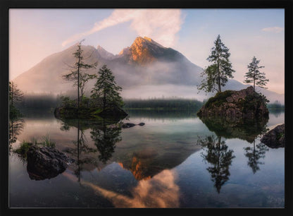 A serene mountain lake at sunrise or sunset, with a prominent peak illuminated by golden light. Small, rocky islands with pine trees dot the calm, clear water, which perfectly reflects the sky, trees, and mountains. A light mist hangs over the water and around the base of the mountains in the background. Decor