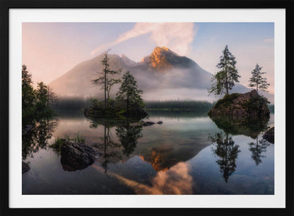 A serene mountain lake at sunrise or sunset, with a prominent peak illuminated by golden light. Small, rocky islands with pine trees dot the calm, clear water, which perfectly reflects the sky, trees, and mountains. A light mist hangs over the water and around the base of the mountains in the background. Decor