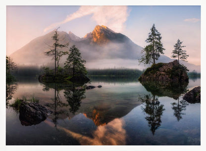 A serene mountain lake at sunrise or sunset, with a prominent peak illuminated by golden light. Small, rocky islands with pine trees dot the calm, clear water, which perfectly reflects the sky, trees, and mountains. A light mist hangs over the water and around the base of the mountains in the background. Decor