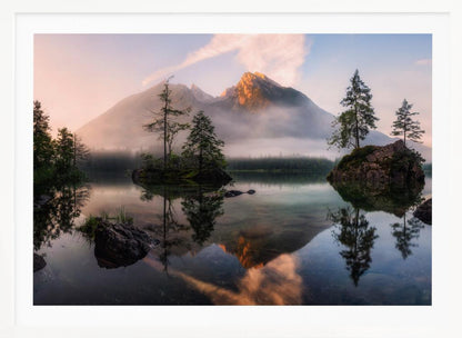 A serene mountain lake at sunrise or sunset, with a prominent peak illuminated by golden light. Small, rocky islands with pine trees dot the calm, clear water, which perfectly reflects the sky, trees, and mountains. A light mist hangs over the water and around the base of the mountains in the background. Decor
