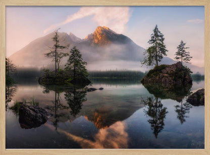 A serene mountain lake at sunrise or sunset, with a prominent peak illuminated by golden light. Small, rocky islands with pine trees dot the calm, clear water, which perfectly reflects the sky, trees, and mountains. A light mist hangs over the water and around the base of the mountains in the background. Decor