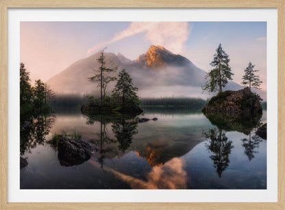 A serene mountain lake at sunrise or sunset, with a prominent peak illuminated by golden light. Small, rocky islands with pine trees dot the calm, clear water, which perfectly reflects the sky, trees, and mountains. A light mist hangs over the water and around the base of the mountains in the background. Decor
