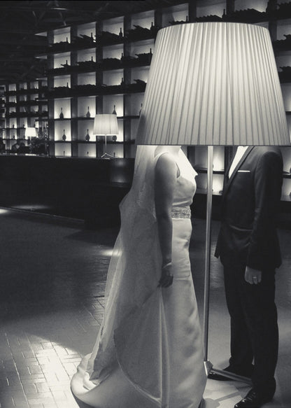 A surreal black and white photograph of a bride and groom whose heads are comically obscured by a large, pleated lampshade from a floor lamp standing between them. They are in a modern, dimly lit room with a wall of illuminated shelves. Poster