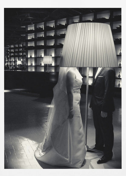 A surreal black and white photograph of a bride and groom whose heads are comically obscured by a large, pleated lampshade from a floor lamp standing between them. They are in a modern, dimly lit room with a wall of illuminated shelves. Poster
