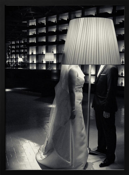 A surreal black and white photograph of a bride and groom whose heads are comically obscured by a large, pleated lampshade from a floor lamp standing between them. They are in a modern, dimly lit room with a wall of illuminated shelves. Poster