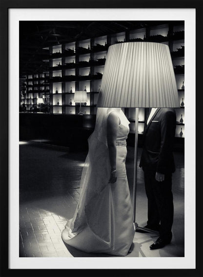 A surreal black and white photograph of a bride and groom whose heads are comically obscured by a large, pleated lampshade from a floor lamp standing between them. They are in a modern, dimly lit room with a wall of illuminated shelves. Poster