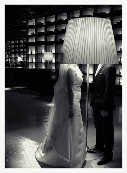 A surreal black and white photograph of a bride and groom whose heads are comically obscured by a large, pleated lampshade from a floor lamp standing between them. They are in a modern, dimly lit room with a wall of illuminated shelves. Poster